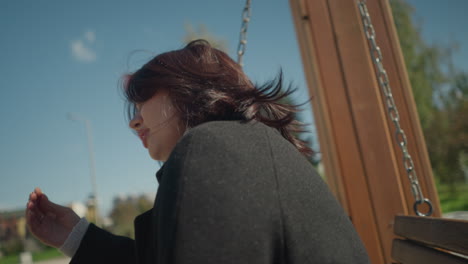 primer plano de una joven sonriendo mientras se balancea al aire libre, su cabello balanceándose con la brisa mientras lo ajusta, rodeado de vegetación en un parque soleado y brillante