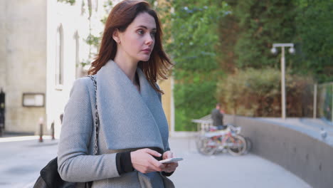 Young-white-adult-woman-standing-on-a-street-in-London-using-her-smartphone,-turns-and-walks-out-of-shot-close-up,-focus-on-foreground