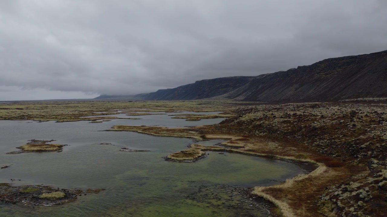 Vådområdepøle og mosdækket vulkansk terræn strækker sig under en barsk højderyg i det sydvestlige Island, med lavvandede kanaler der krydser det klippefyldte terræn under en droneglidning.