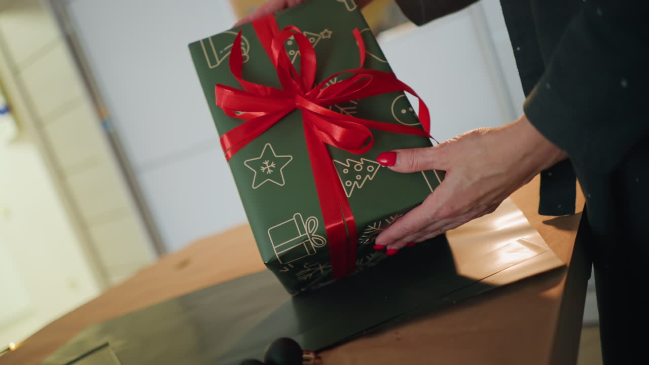 Close-up de mãos embrulhando presente de Natal em mesa de madeira com papel estampado de estrelas verdes e fita vermelha, dobradura e amarração cuidadosas, acabamento de laço impecável, cena aconchegante de cozinha interna, preparação para o feriado