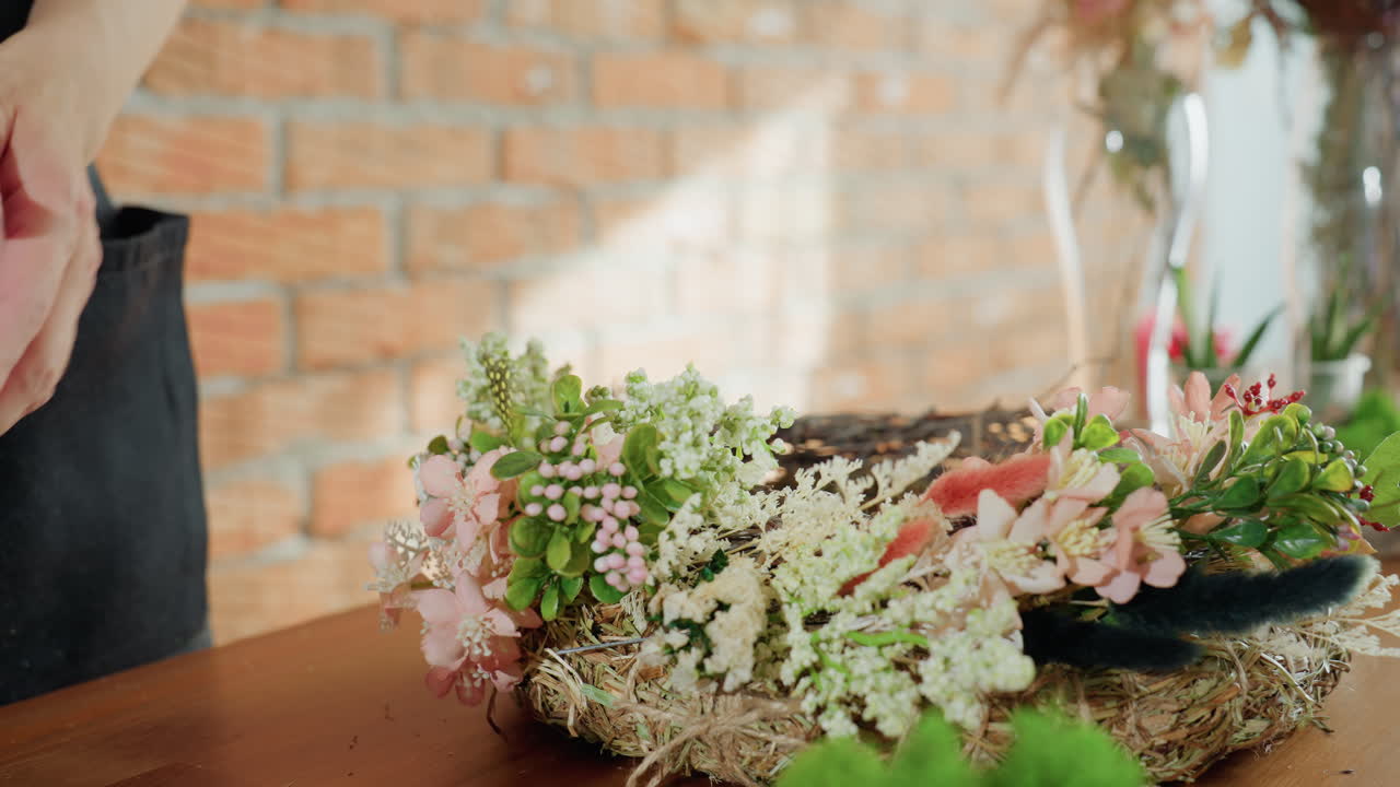 Female florist with red nails adjusting handmade floral wreath with delicate flowers and greenery on rustic straw base, close-up view of hands working with natural materials in creative workshop atmosphere