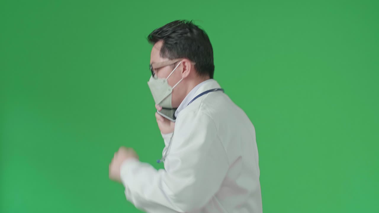 The Side View Of Asian Doctor With Stethoscope Wearing Mask, Glasses, And White Coat Talking On Mobile Phone While Running On Green Screen Chroma Key.
