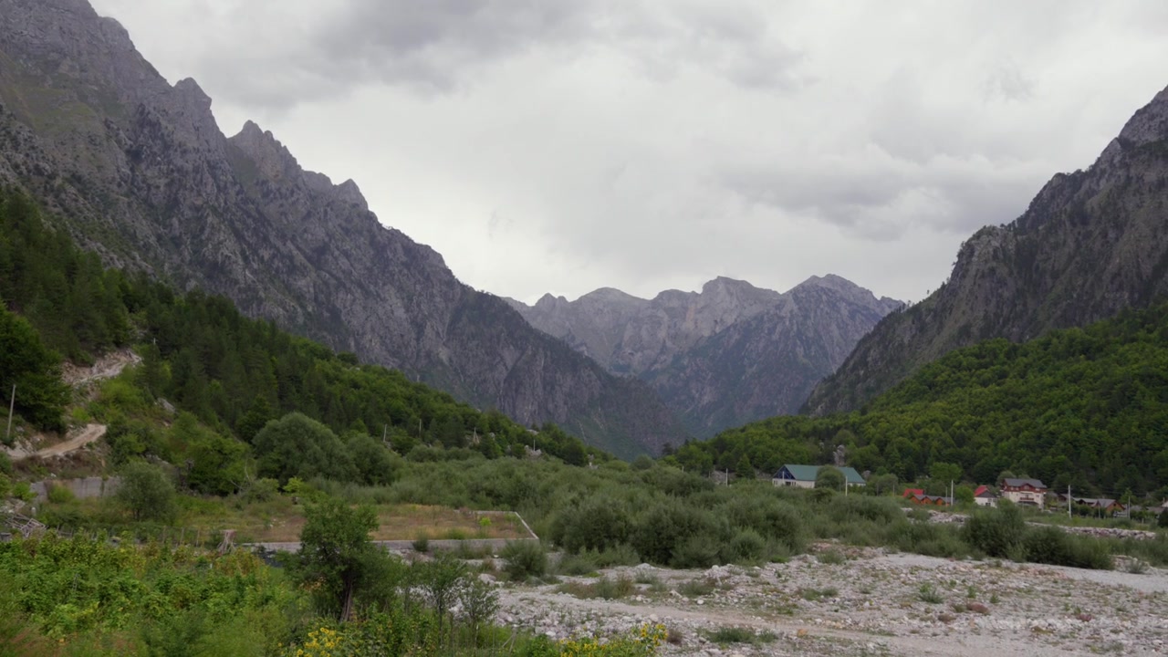 Vidstrakte, steinete fjell reiser seg over frodige grønne skoger og spredte hus under en skyet himmel i Albanias Valbona-dal.