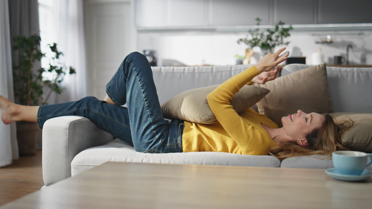 Carefree woman texting telephone message laying sofa home. Happy woman chatting