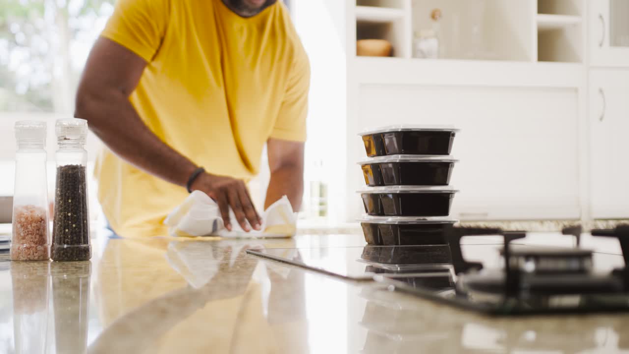 Homem negro limpando a bancada de pedra da cozinha com pano branco, preparando recipientes para refeições