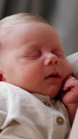 tiny caucasian infant sleeping in mom's arms holding little fist at his cheek. mom touches baby nose with the finger tenderly. close up. vertical video