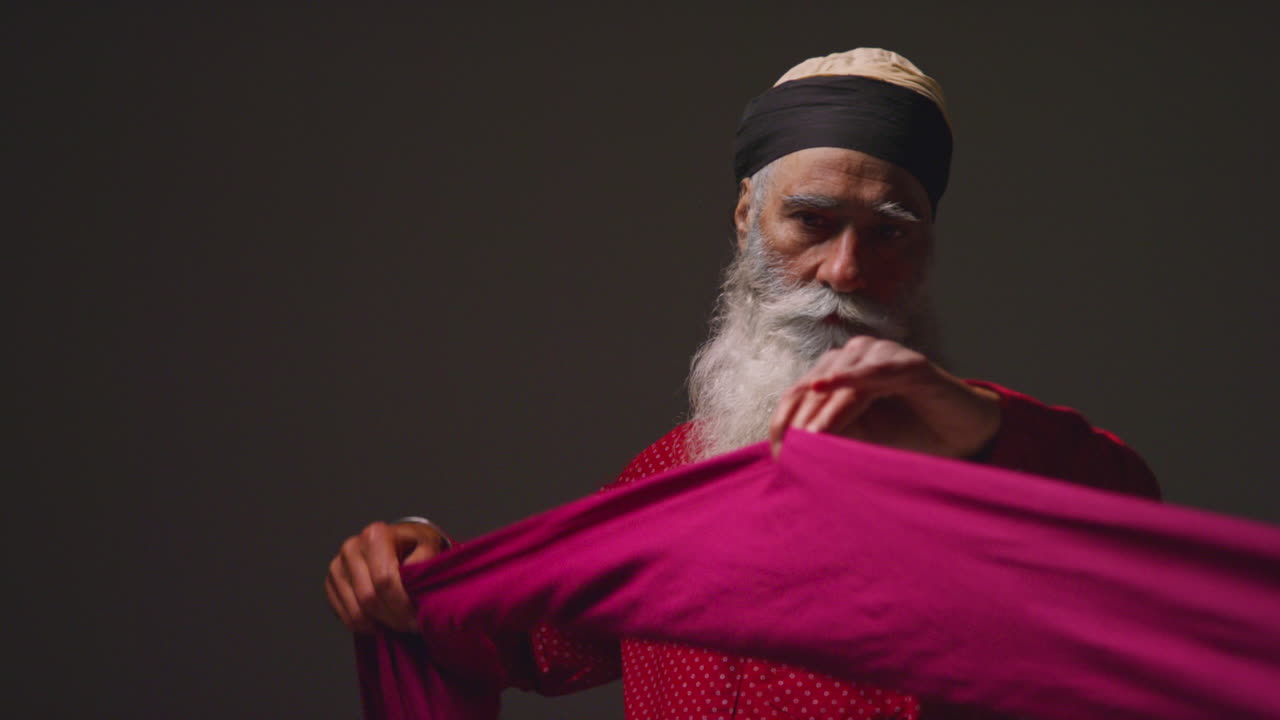 Low Key Studio Lighting Shot Of Senior Sikh Man Folding Fabric For Turban Against Plain Dark Background 3
