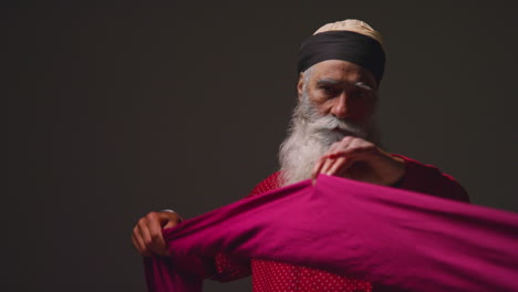 Fotografía-De-Estudio-Con-Iluminación-Tenue-De-Un-Hombre-Sikh-Mayor-Doblando-Tela-Para-Un-Turbante-Contra-Un-Fondo-Oscuro-Liso-3