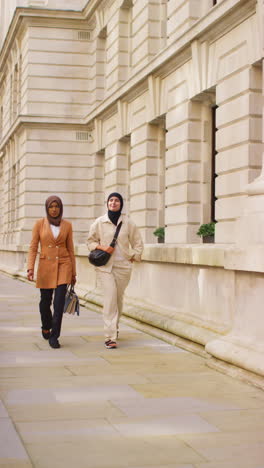 vertical video of two muslim businesswomen wearing hijabs with modern business suits walking to work past city office buildings