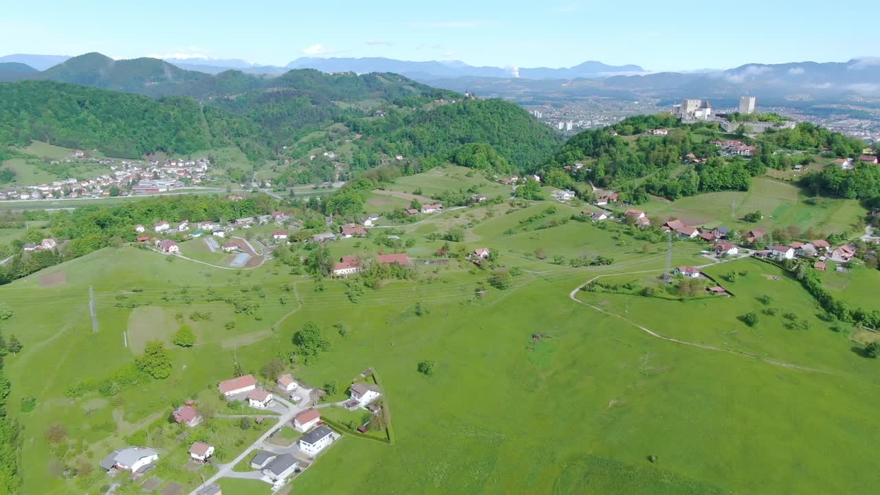 Hilly lush green landscape with farms, isolated houses and a town in the valley connected by winding roads on a sunny day. Aerial drone shot. Kočevje - Slovenia