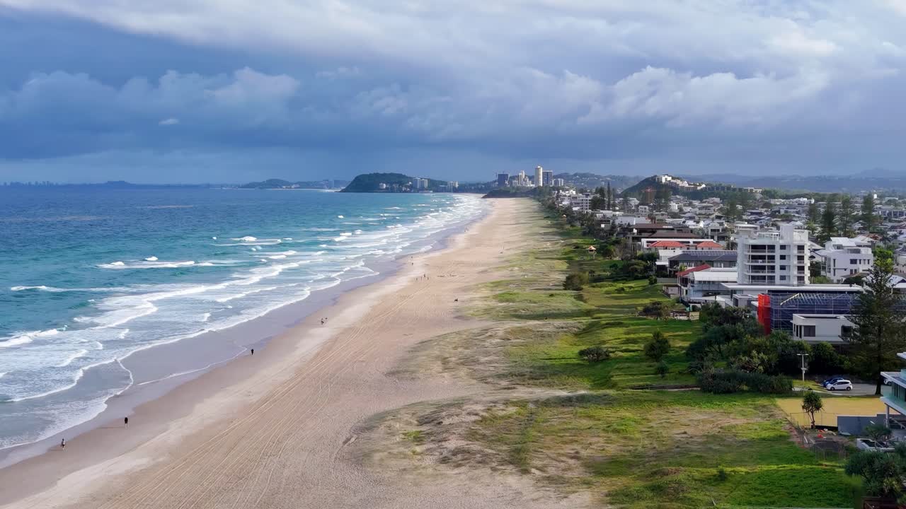 Aerial footage showcasing a sandy beach, ocean waves, and beachfront buildings under a cloudy sky.