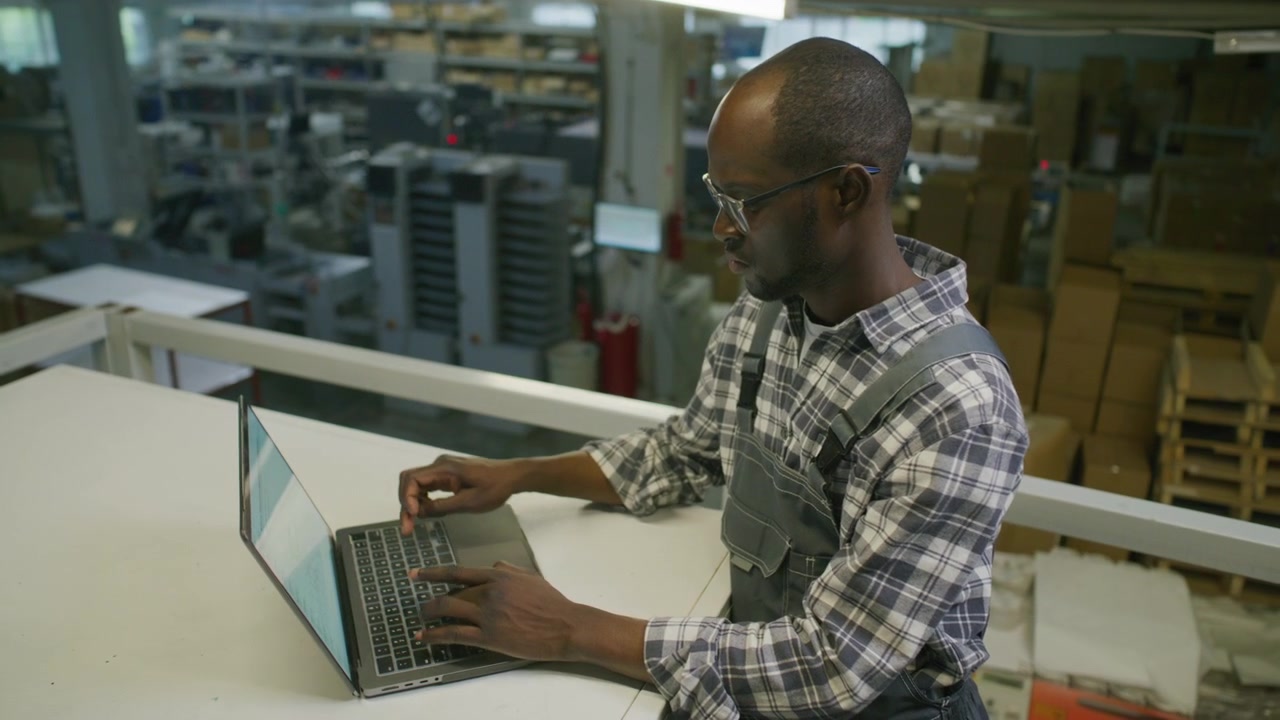 Black Technician Working on Laptop in Industrial Warehouse of Print Center