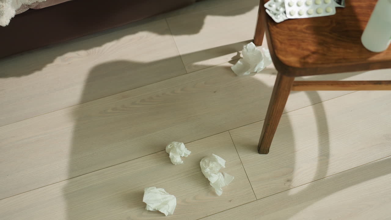 바닥에 있는 사용한 휴지와 알약, 의자 그림자를 드리우는 아침 햇살, 코 스프레이 병 옆에 있는 블리스터 포장; 흩어진 휴지, 조용한 정적, 미묘한 따뜻함이 있는 가정 내 회복 장면