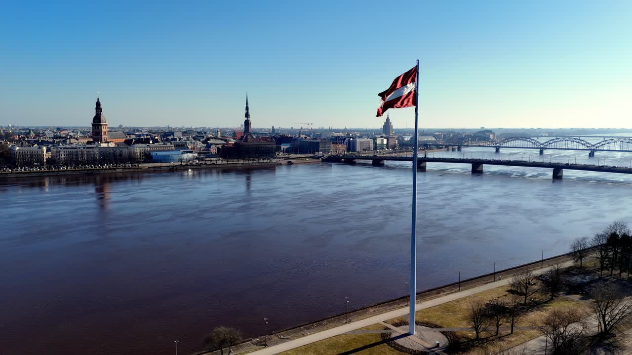 En flagstang står foran en by og et vandområde, Daugava-floden