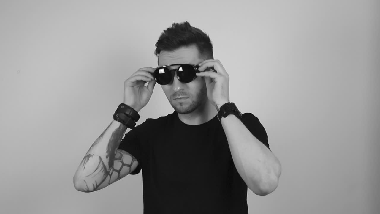 Grayscale footage of a tattooed man with short hair and beard putting on vintage retro steampunk sunglasses in slow motion, watching forward and nodding with confidence while crossing his arms.