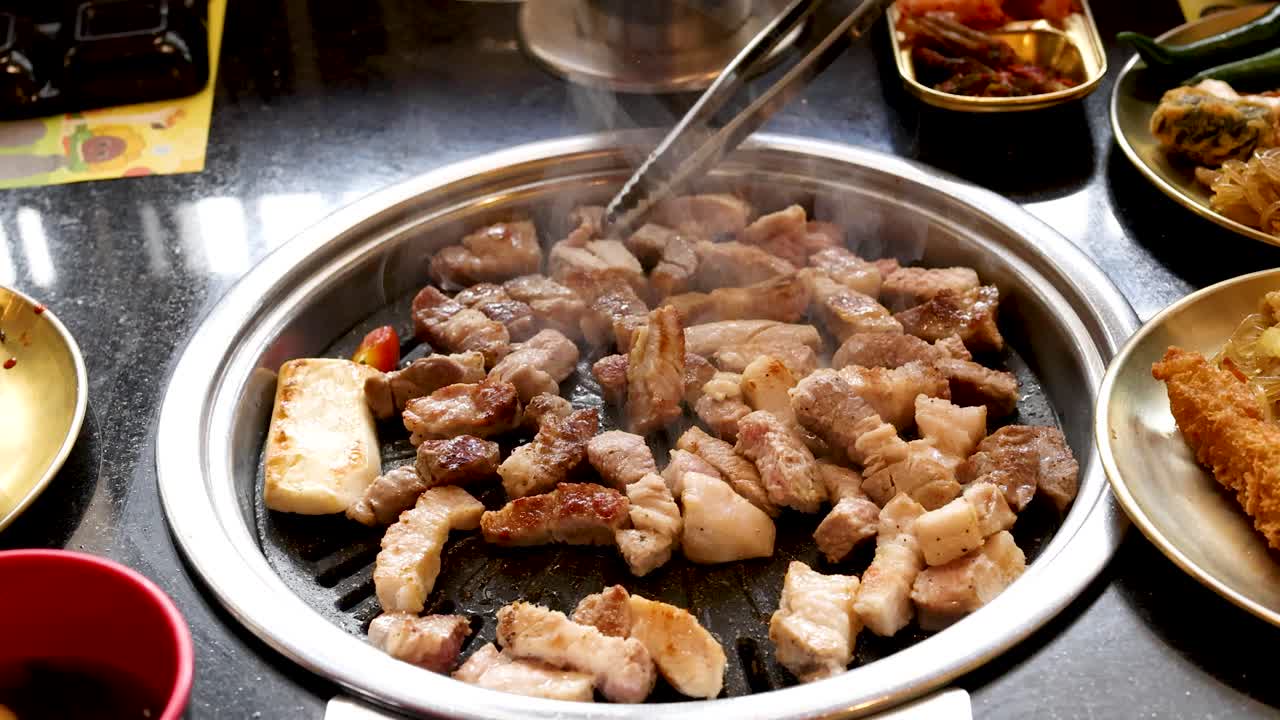 Hands grill pork belly on tabletop grill, surrounded by side dishes, with natural indoor lighting