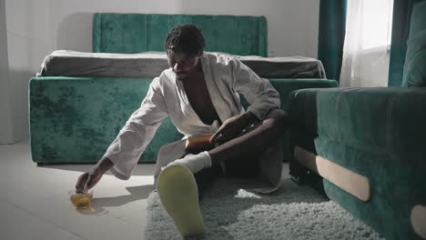 man in bathrobe sitting on floor near bed, stretching and reaching for his tea cup on soft rug, relaxed morning routine in cozy bedroom with green velvet bed and natural light