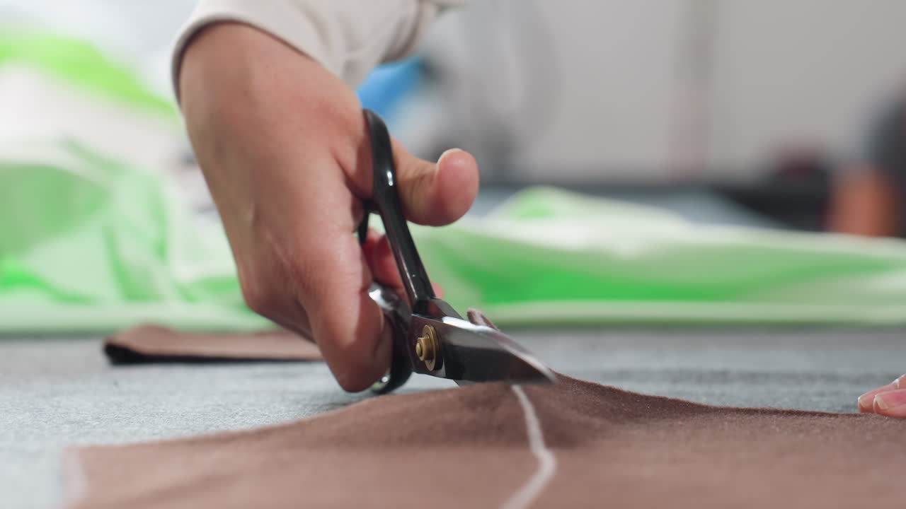 close up of seamstress hand firmly holding scissors cutting brown fabric along white chalk line on work table, precision cutting process in tailoring workshop, focus on accurate textile cutting