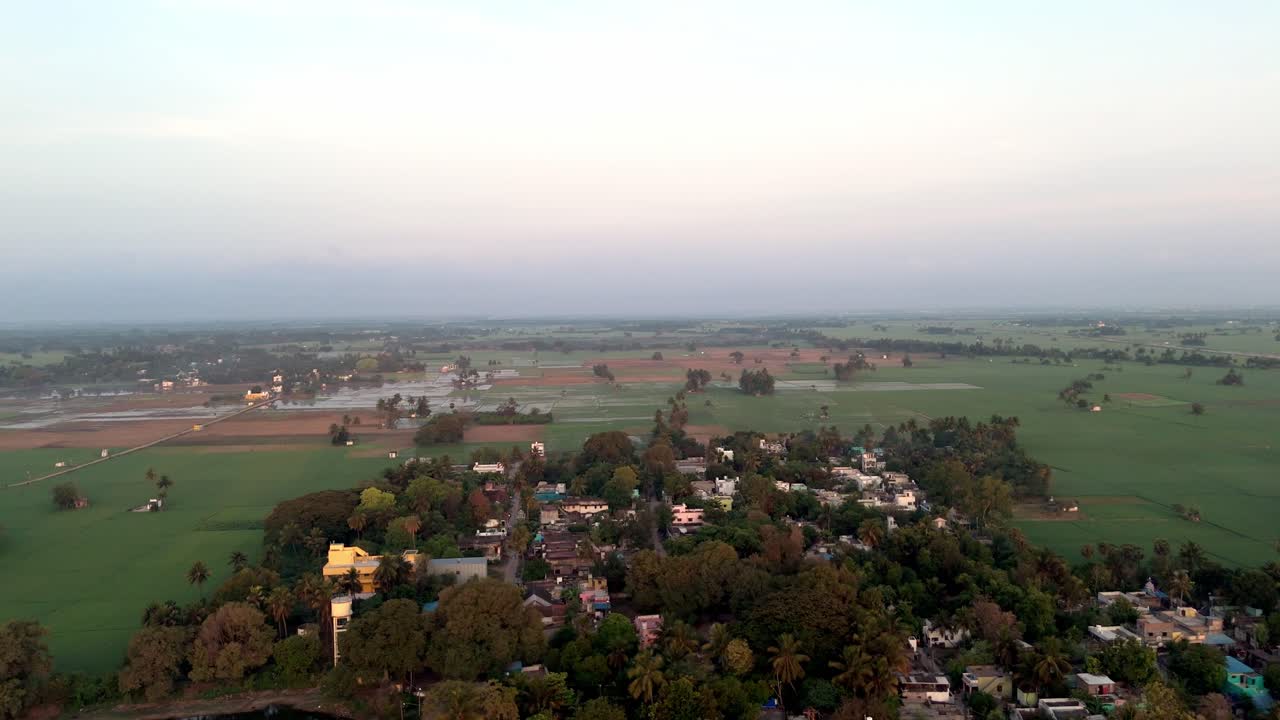 Un'ampia veduta aerea cattura un affascinante villaggio rurale immerso tra rigogliosi terreni agricoli verdi. Il primo piano mostra un gruppo di case e alberi abbondanti, che lasciano il posto a vasti campi agricoli.