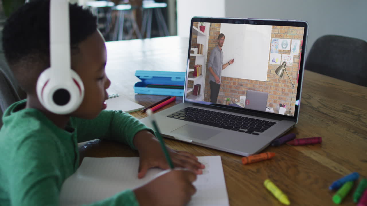African american boy wearing headphones having a video call on laptop while doing homework at home