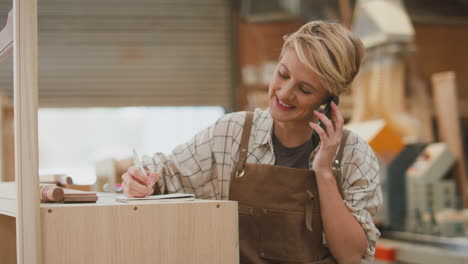 Female-Apprentice-Carpenter-Working-In-Furniture-Workshop-On-Phone-Call-Making-Notes