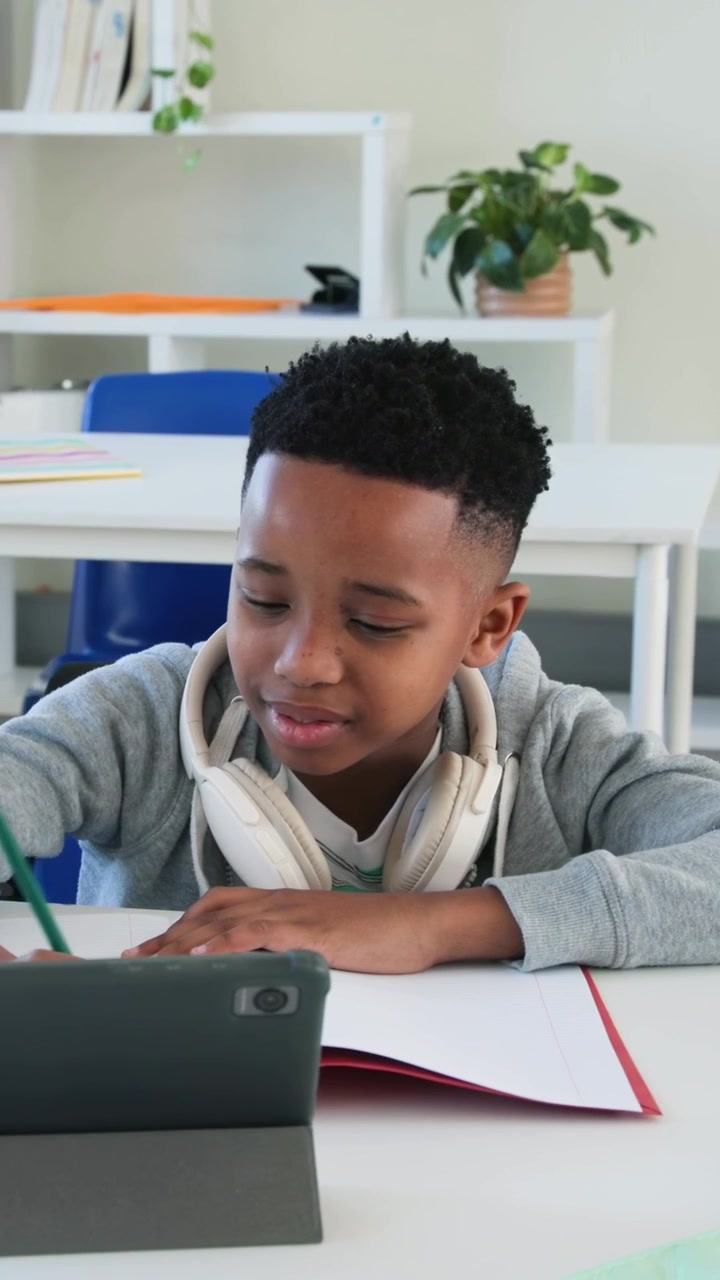 Vertical video: Adjusting student in hoodie handling phones at desk using tablet and writing notes