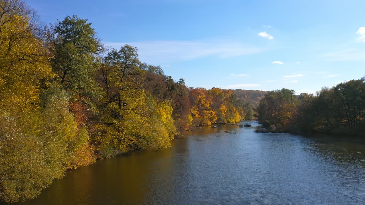 Beautiful autumn scenery. Calm blue river surrounded with yellow, red and orange tree under blue sky. Panoramic view.