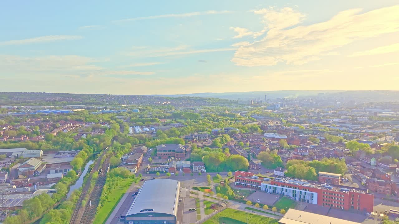 High aerial view reveals Don Valley Bowl's arenas, train tracks, and surrounding green suburbs bathed in warm sunlight with distant skyline and rolling hills across Sheffield, slow motion