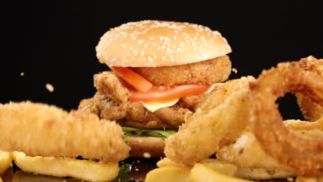 golden onion rings and french fries fall in slow motion beside a fried chicken burger on a glossy black surface, under bright studio lighting