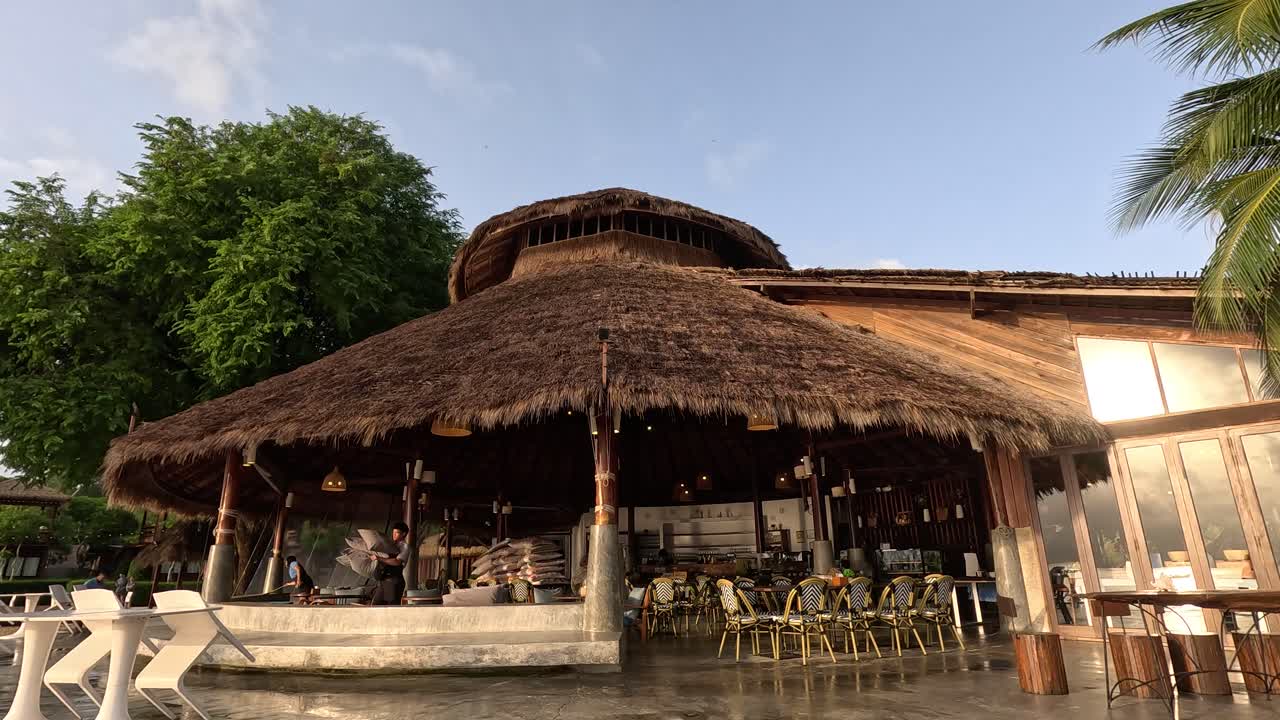 Camera slowly pans down to reveal a thatched-roof beach bar surrounded by palm trees and lush greenery in soft natural daylight