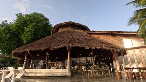 camera slowly pans down to reveal a thatched-roof beach bar surrounded by palm trees and lush greenery in soft natural daylight