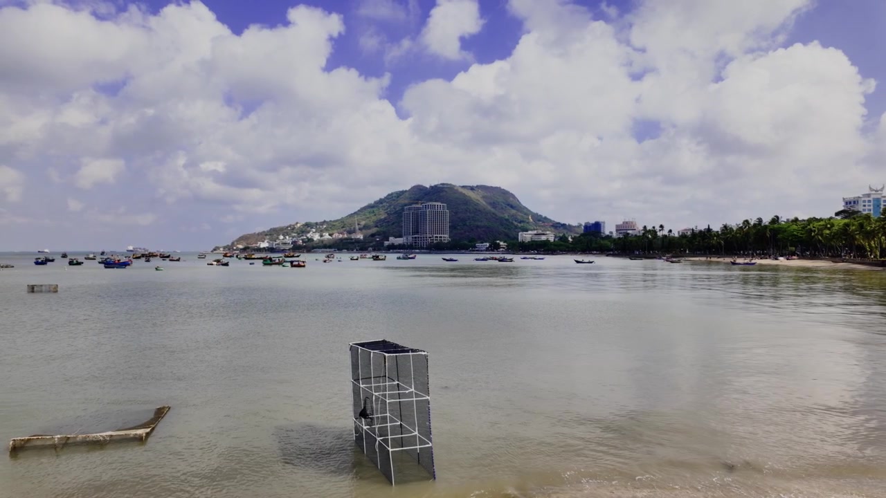 Bai Truoc em Vung Tau oferece uma vista da praia e do mar com vários barcos. A paisagem inclui uma colina e edifícios sob um céu limpo. As pessoas aproveitam a área e o mar.