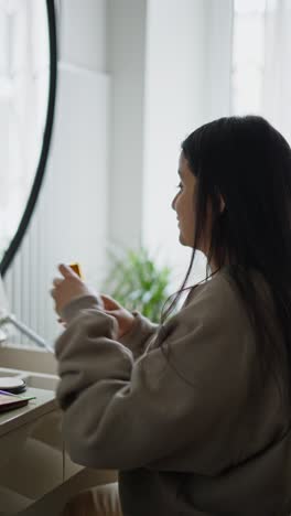 Vertical-video-of-a-confident-brunette-girl-in-a-gray-sweater-doing-Makeup-and-trying-the-smell-of-new-perfume-in-front-of-a-large-mirror-in-a-modern-apartment