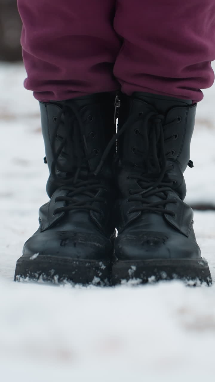 primer plano de piernas con botas negras mientras la persona salta de izquierda a derecha en el suelo cubierto de nieve, las botas dejan ligeras huellas en la nieve fresca, con aire frío de invierno y fondo borroso