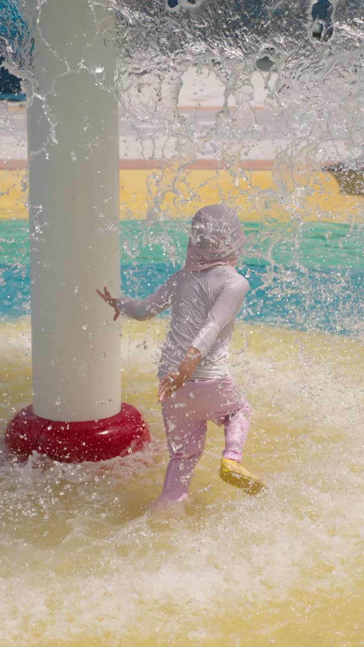 Vertical slow motion shot capturing a joyful young girl running and playing underneath a splashing water fountain, having fun at an outdoor summer water playground attraction