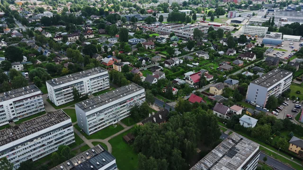 Orbiting drone view over Darzciems in Riga, Latvia, with panel apartment blocks beside houses and gardens, and a newly paved asphalt road running through the neighborhood