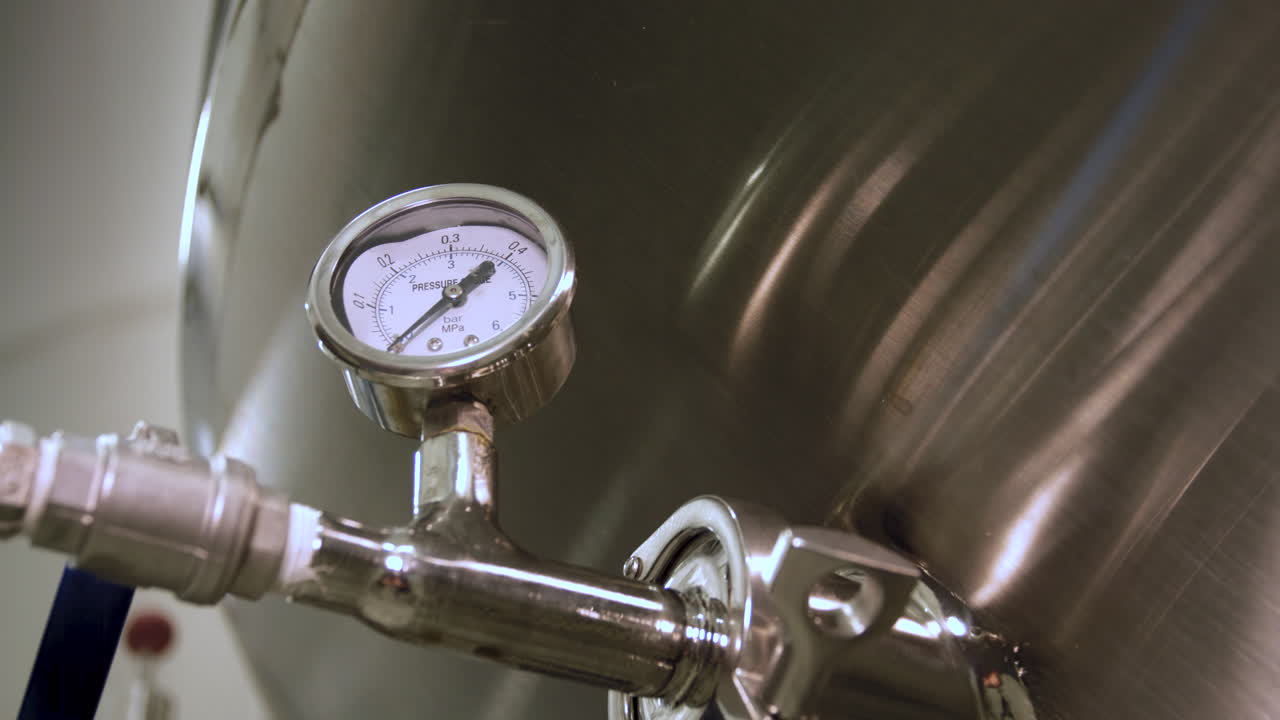 Rack focus from a close-up of a pressure gauge mounted on a stainless steel fermentation tank in a brewery to a blurry view