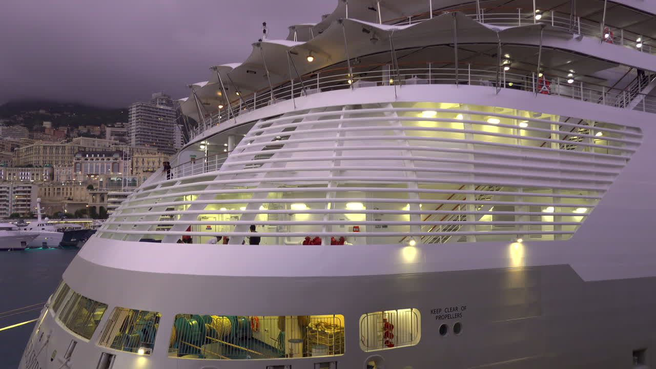 La Condamine, Monaco - July 4, 2025: Evening view of a cruise ship docked in Monaco, with illuminated decks and the city skyline in the background under a cloudy sky