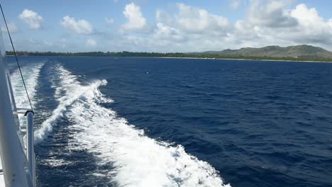 Panoramic-view-from-side-of-boat-in-motion,-making-choppy-waves,-looking-back-at-land-in-Guam