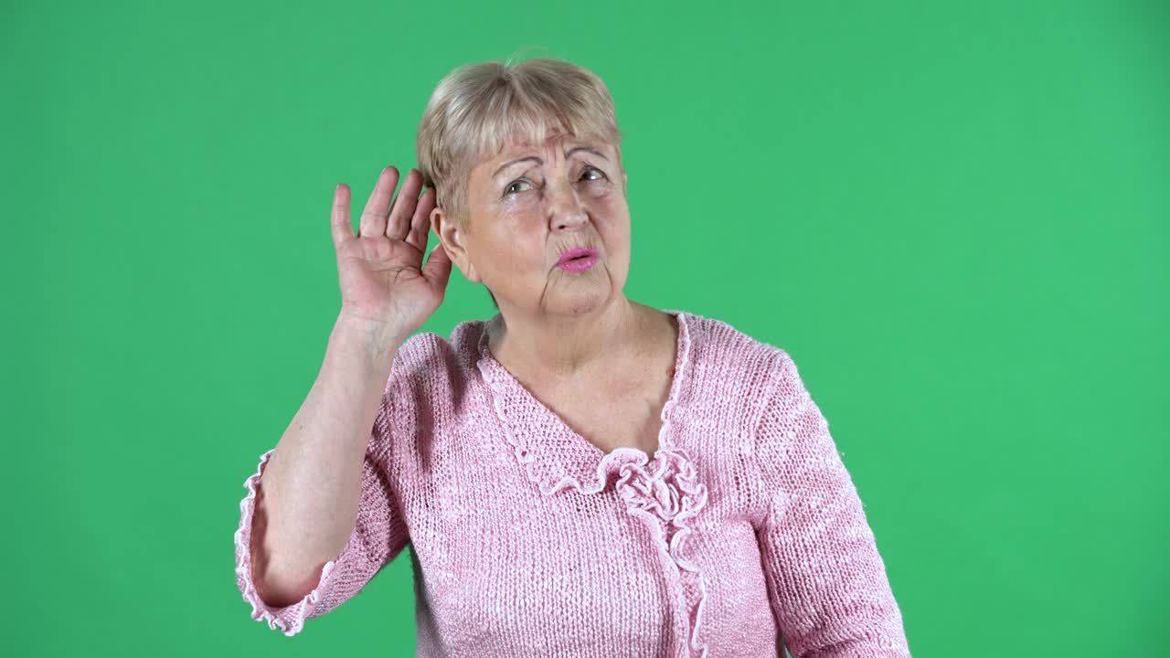 retrato mujer anciana sosteniendo la mano cerca de la oreja tratando de escuchar noticias interesantes expresando concepto de comunicación y chismes. abuela de cabello gris en un suéter rosa en la pantalla verde en el estudio. cerca