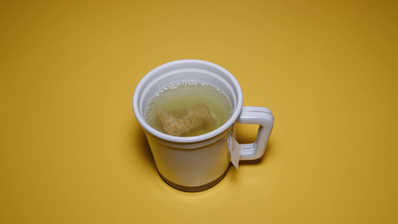 Una ripresa a mezzo busto di una tazza da tè con una bustina all'interno, mentre viene versata acqua calda da un thermos, facendo galleggiare la bustina. La scena evoca un'atmosfera calda e pacifica