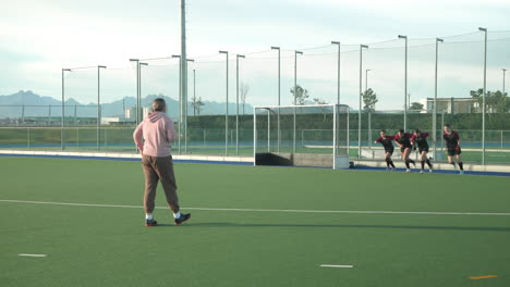female hockey team training on field, coach observing players' warm-up routine