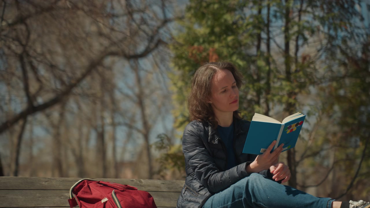 Close up of woman reading book on wooden bench with red bag nearby in park setting. Subject holds open blue covered volume maintaining focus throughout calm outdoor scene with trees