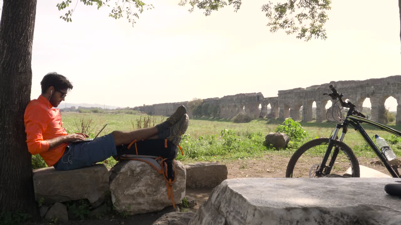 Ciclista con la bici seduto su una roccia sotto un albero che scrive al portatile davanti a un antico acquedotto romano. Giovane uomo attraente e atletico con abbigliamento sportivo arancione e zaino nel Parco degli Acquedotti a Roma
