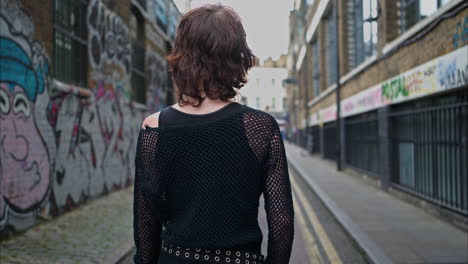 rear view outdoor fashion shot of young alternative style woman walking past graffiti covered walls in london city street uk in real time