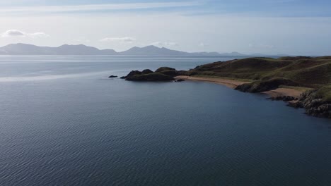 Aerial-view-across-idyllic-Ynys-Llanddwyn-island-with-hazy-Snowdonia-mountain-range-across-shimmering-Irish-sea-at-sunrise,-Zoom-out