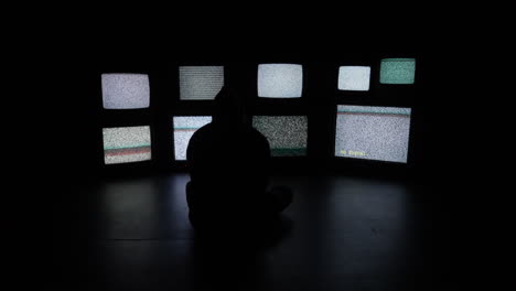 an anxious and isolated person sits in front of a wall of white noise filled televisions media screens in a dark room