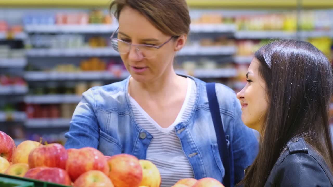 Engaging Moment in a Grocery Store: A Woman Analyzes Apples While Shopping with a Friend, Reflecting the Joy of Food Selection and Connection