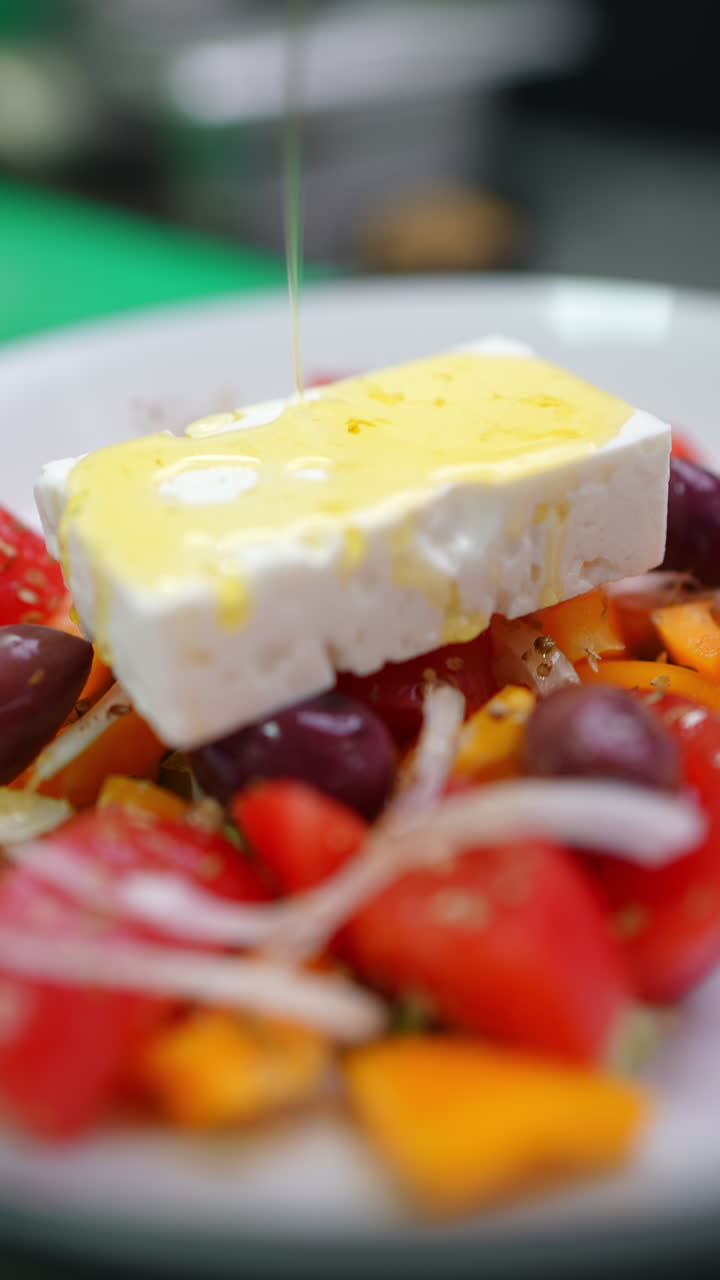 Vertical footage Feta cheese is placed on a traditional greek salad with tomatoes, cucumbers, onions, olives, peppers and olive oil being poured on top