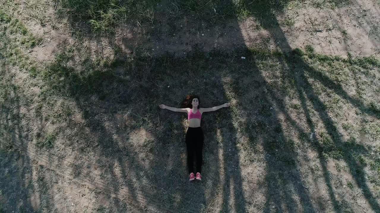 나무 옆에 팔을 벌리고 바닥에 누워 있는 젊은 갈색 머리 여성의 드론 샷